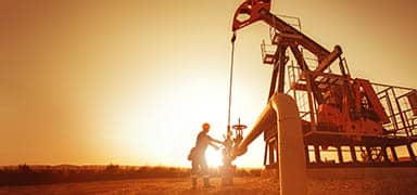 A worker operates equipment at an oil pumpjack site during sunset, with warm sunlight illuminating the machinery and landscape.