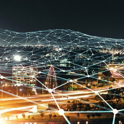 A cityscape at night with bright lights, busy roads, and buildings, overlaid with glowing blue lines and dots representing a digital network or connectivity.