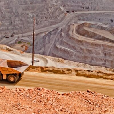 A large dump truck drives along a dirt road at the edge of an expansive open-pit mine, with terraced layers and winding paths visible in the background.