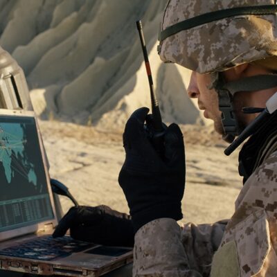 A soldier in camouflage uniform uses a walkie-talkie while looking at a laptop displaying a world map with connecting lines, suggesting military communications or operations in a desert environment.