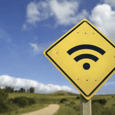 A yellow road sign with a black Wi-Fi symbol stands by a dirt road in a rural landscape, with green fields, trees, and a blue sky with scattered clouds in the background.