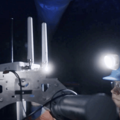 A person wearing a blue hard hat and headlamp operates or observes a machine with antennas and robotic arms in a dimly lit, industrial or underground environment.