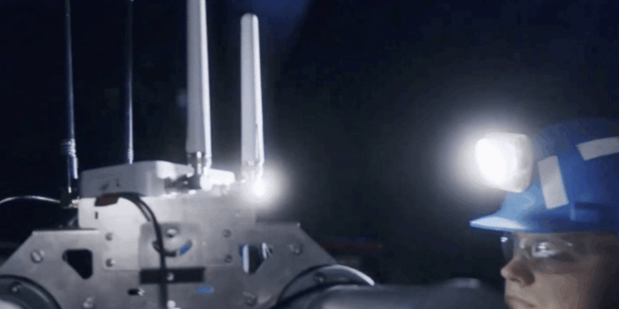 A person wearing a blue hard hat and headlamp operates or observes a machine with antennas and robotic arms in a dimly lit, industrial or underground environment.