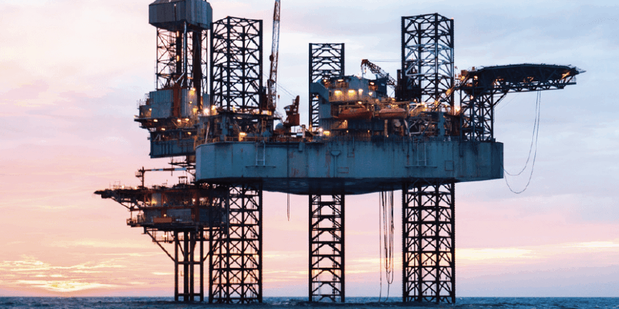An offshore oil rig stands elevated above the ocean water at sunset, with its large platform, cranes, and support structures visible against a colorful sky.