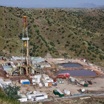 An aerial view of a drilling site in a hilly, green landscape. The site has a tall drilling rig, several large water or mud pits, and numerous white containers and vehicles scattered around the area.