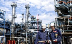 Two workers in protective gear and helmets inspect a clipboard at an industrial oil refinery with tall metal towers and pipes against a partly cloudy sky.