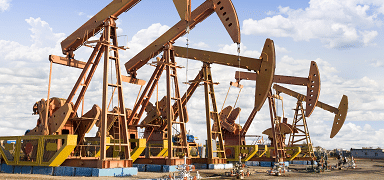 Several oil pumpjacks operate in a row on an oil field under a partly cloudy sky, extracting oil from underground. The scene is industrial, with metal equipment and a flat landscape visible.