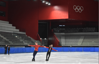 Two figure skaters perform on an indoor ice rink with empty seats and the Olympic rings displayed on a red wall in the background. Other skaters are visible practicing nearby.