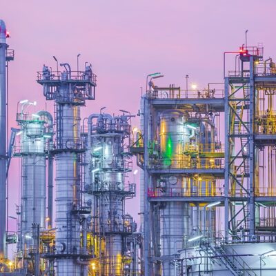 Industrial refinery towers and piping illuminated by lights at dusk, with a pink and purple sky in the background. The structures are complex, featuring ladders, platforms, and various cylindrical tanks.