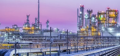 A large industrial oil refinery with numerous pipes, towers, and storage tanks, illuminated by bright lights against a purple and pink dusk sky.