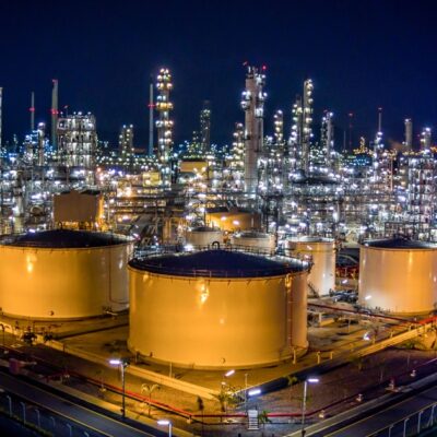 A brightly lit oil refinery at night, featuring large cylindrical storage tanks and numerous tall industrial towers with lights, pipes, and platforms. The surrounding area is illuminated with streetlights.