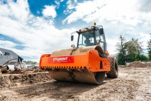 An orange road roller with "STORENT" on the front is parked on a construction site with dirt and debris, under a partly cloudy sky. Trees and buildings are visible in the background.