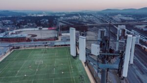 Aerial view of a green soccer field near a railway, with a large cell tower in the foreground and an industrial cityscape in the background at dusk.