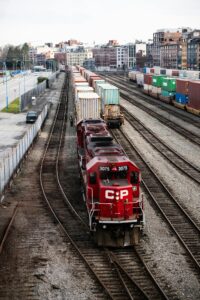 A red CP freight train engine pulls container cars along multiple railway tracks in a rail yard, with city buildings and parked containers visible in the background.