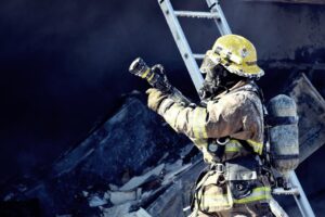 A firefighter in full protective gear and helmet holds a hose, aiming it upwards near a ladder, with smoke and debris visible in the background.