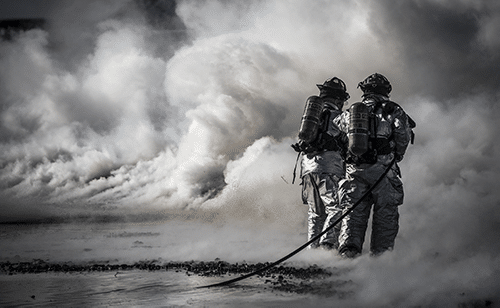 Two firefighters in protective gear and oxygen tanks stand amid thick smoke, holding a hose and facing away from the camera, apparently fighting a fire in a smoky outdoor environment.