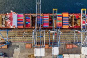 Aerial view of a cargo ship docked at a port, loaded with colorful shipping containers. Large cranes are positioned over the vessel, and the dock below shows vehicles and storage areas.