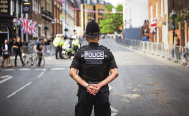 A police officer stands in the middle of a street with hands behind his back, facing away. Barricades line the road, with people, bicycles, and British flags visible in the background.