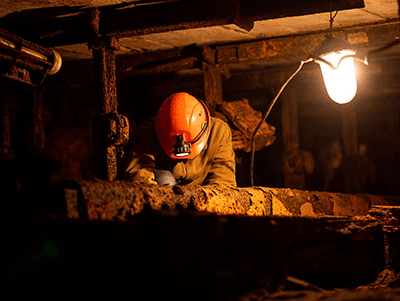 A miner wearing an orange helmet and headlamp works underground, illuminated by a single hanging light amid rusty beams and machinery.