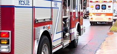A fire truck and an ambulance are parked on a wet road, suggesting an emergency response scene. The vehicles’ lights are visible, and the road and nearby grass appear damp from recent rain.