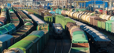 A large rail yard filled with numerous freight trains, with green, brown, and white cargo cars arranged on parallel tracks, set against an industrial backdrop.