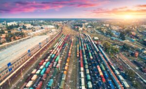 Aerial view of a large railway yard with many colorful freight trains and tracks at sunset, with network-like digital graphics overlaying the scene, symbolizing technology and connectivity in transportation.