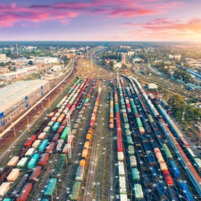 Aerial view of a large railway yard with many colorful freight trains and tracks at sunset, with network-like digital graphics overlaying the scene, symbolizing technology and connectivity in transportation.
