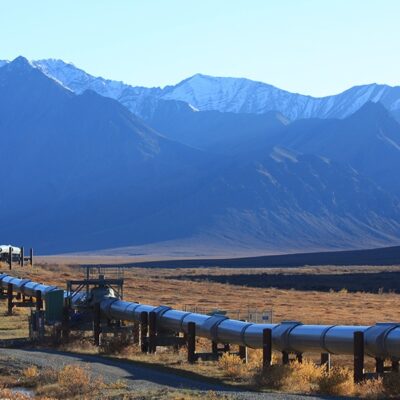 A large metal pipeline stretches across a flat, grassy landscape with rugged mountains in the background under a clear blue sky.