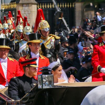 A royal wedding procession with a couple in a horse-drawn carriage, escorted by guards in red uniforms and helmets, passing by a large crowd behind barriers. The bride waves and the groom sits beside her.