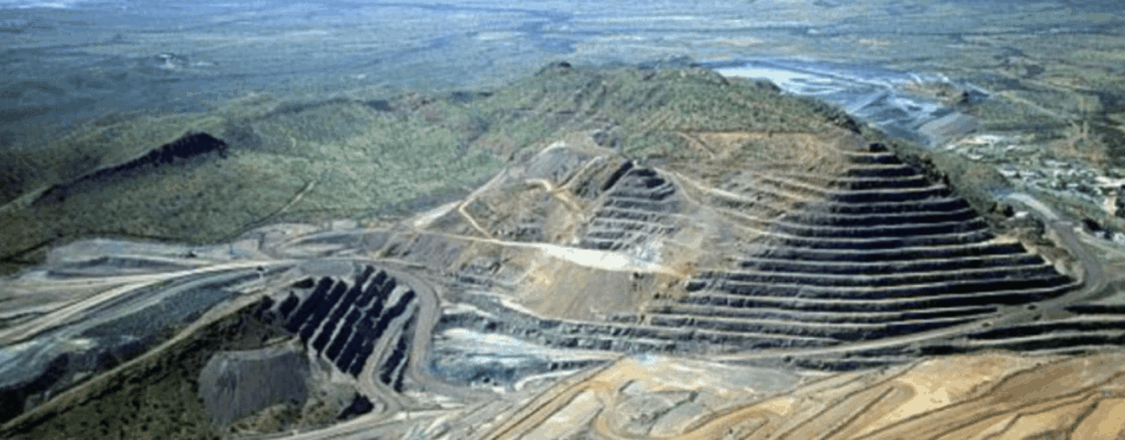 Aerial view of a large open-pit mine with terraced, stepped sides carved into a hillside. Surrounding areas show sparse vegetation and winding roads amid the mining activity.