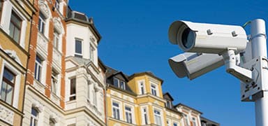 A pair of security cameras mounted on a white pole overlook a row of colorful, ornate residential buildings under a clear blue sky.