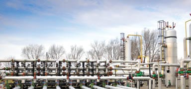 An industrial facility with multiple large pipes, valves, and storage tanks outdoors, set against a background of leafless trees and a partly cloudy sky.