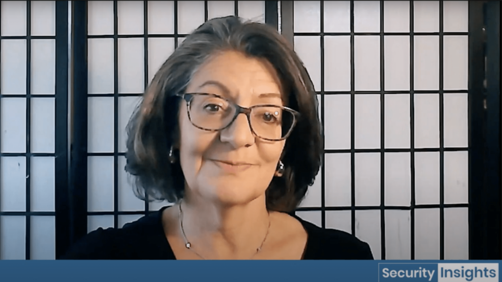 A woman with shoulder-length brown hair and glasses smiles at the camera, seated in front of a black and white grid-patterned screen. Text in the lower right corner reads "Security Insights.