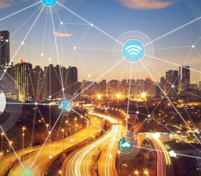 A city skyline at sunset with highways and light trails, overlaid with digital network icons representing smart technology and connectivity, illustrating the concept of a smart city and Internet of Things (IoT).