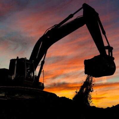 A silhouette of an excavator scooping and dropping dirt, set against a vibrant sunset sky with orange, pink, and blue clouds.