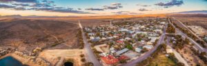Aerial view of a small rural town surrounded by dry, open land and mountains under a blue sky with scattered clouds at sunset. Houses, roads, and some trees are visible in the town.