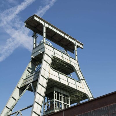 A metal industrial mining tower rises above a brick building under a clear blue sky with white streaks of clouds.