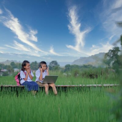 Two young girls in school uniforms sit on a wooden bench in a lush green field, using a laptop together, with blue sky and white clouds in the background. One girl has a pink backpack beside her.