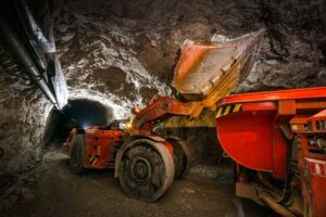 An orange excavator loads rock into a dump truck inside an underground mining tunnel, with rugged rock walls and construction equipment visible.