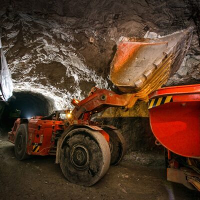 An orange excavator loads rock into a dump truck inside an underground mining tunnel, with rugged rock walls and construction equipment visible.