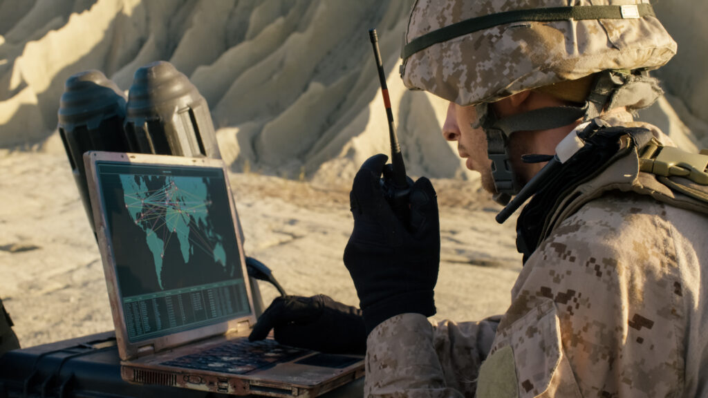 A soldier in camouflage uniform and helmet uses a radio while looking at a laptop displaying a digital map with colored lines, sitting outdoors near tan, rocky terrain.