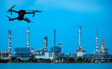 A drone flies in the foreground with an industrial refinery complex and tall chimneys illuminated in the background at dusk.