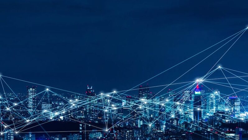 A city skyline at night with illuminated buildings and white lines connecting points across the sky, representing a digital network or smart city technology.