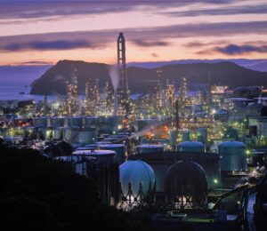 A large industrial oil refinery with storage tanks and spherical gas containers, illuminated by bright lights at dusk, set against hills and a cloudy sky near the coastline.