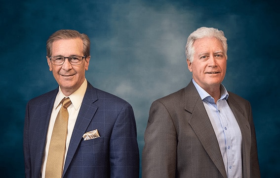 Two older men in suits stand side by side against a blue, textured studio backdrop. One man wears glasses, a beige tie, and a pocket square; the other man has white hair and wears an open-collared shirt.