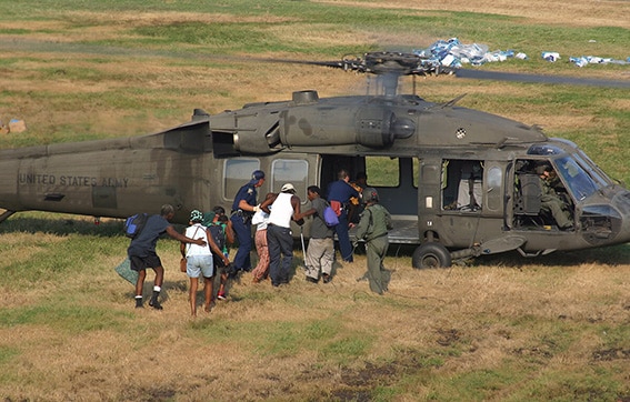 A group of people boards a United States Army helicopter on a grassy field, assisted by uniformed personnel. Supplies are visible scattered in the background.