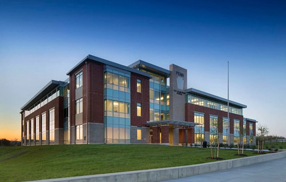 A modern three-story building with large glass windows and red brick walls, set on a grassy hill at sunset, with a clear blue sky in the background.