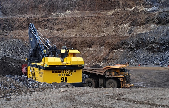A large yellow mining excavator loads rocks into a dump truck in an open-pit mine. The excavator is labeled "IRON ORE COMPANY OF CANADA 98" and is surrounded by rocky terrain.
