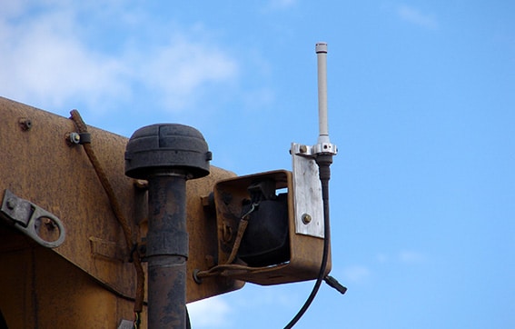 A close-up of an industrial machine part with a small antenna mounted on a metal bracket, a black cylindrical component, and weathered yellow metal against a blue sky with some clouds.