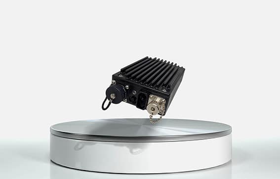 A black electronic device with a ribbed surface and various connectors is suspended above a round, metallic display pedestal against a plain white background.